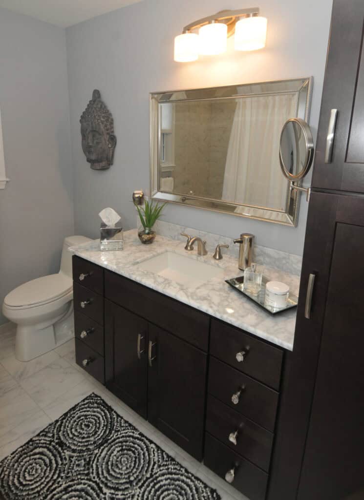 Modern bathroom with a marble countertop, dark wood cabinets, large mirror, wall-mounted Buddha sculpture, toilet, and a tray with cups and soap on the sink. A patterned rug is on the marble floor.