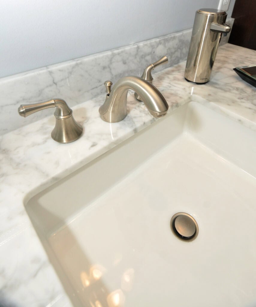 Close-up of a modern bathroom sink with a brushed nickel faucet and handles on a white marble countertop. A soap dispenser is placed near the faucet.