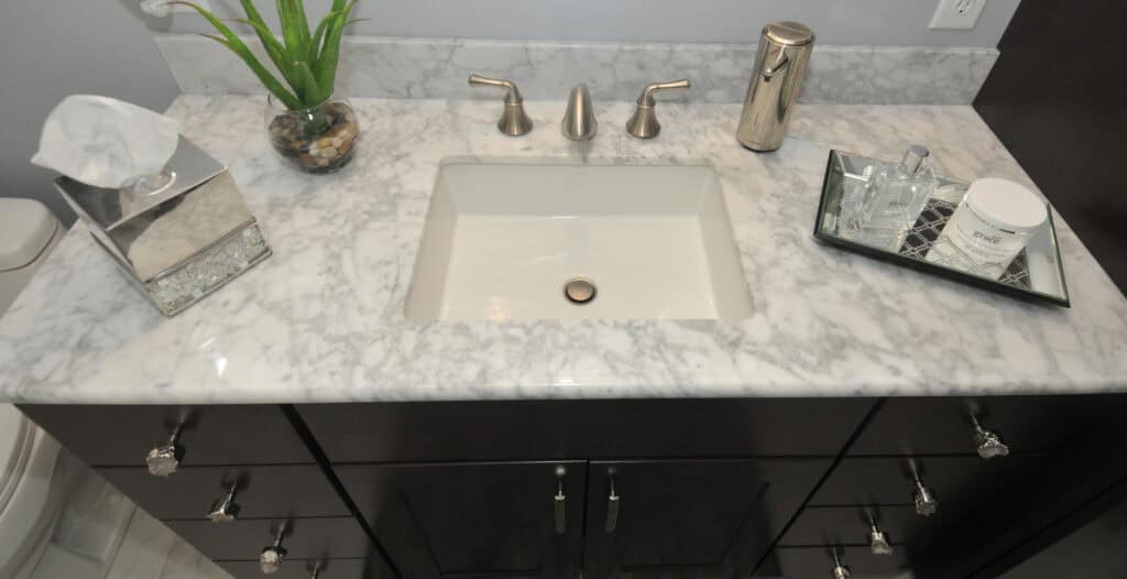 A marble bathroom countertop with a square sink, silver faucet fixtures, tissue box, soap dispenser, small plant, and a tray holding toiletries and a candle. Dark cabinets are visible below the counter.