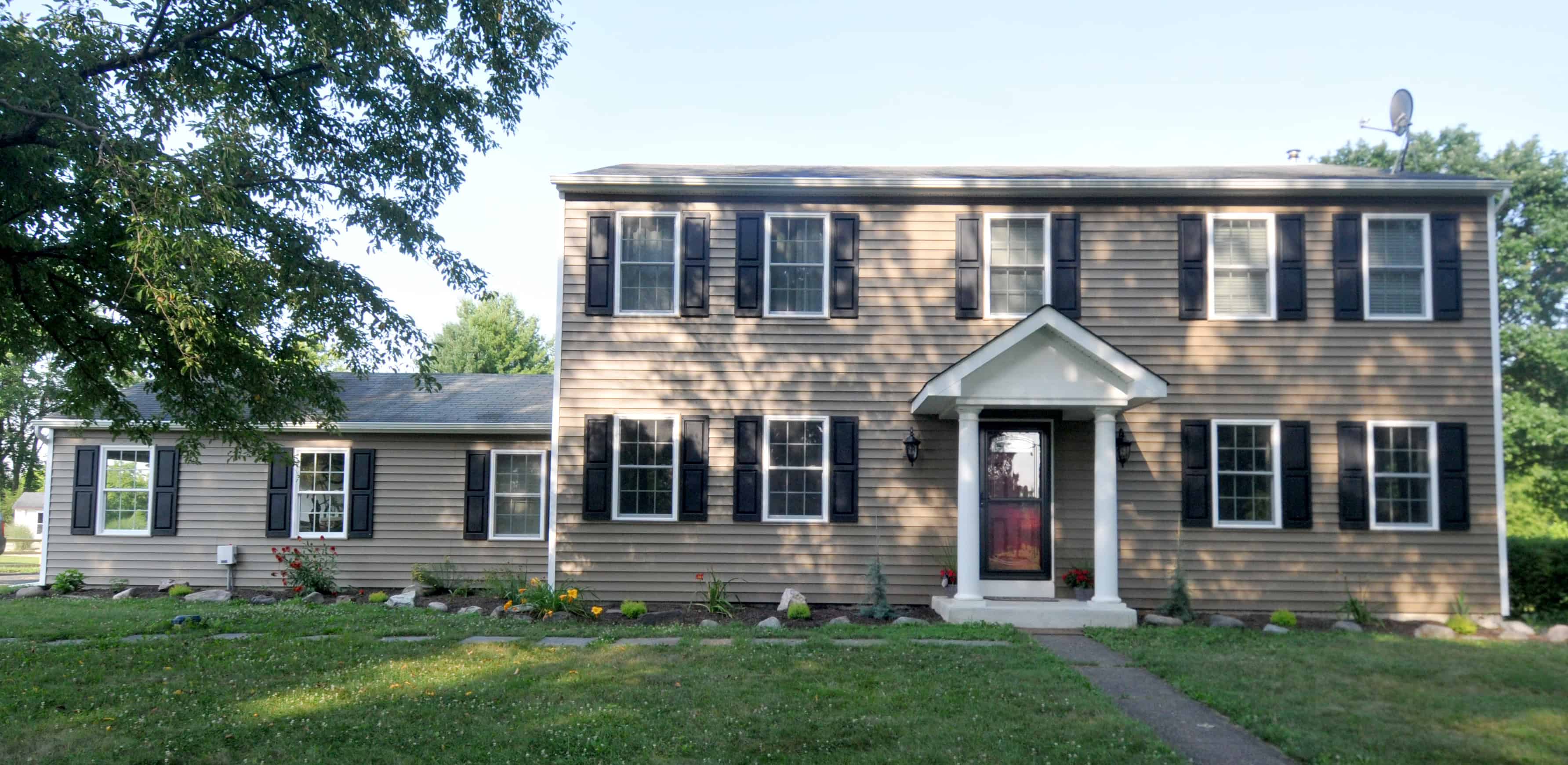 A two-story beige house with black shutters, a red front door, and a small portico, surrounded by grass, a tree, and some small plants, under a clear sky.