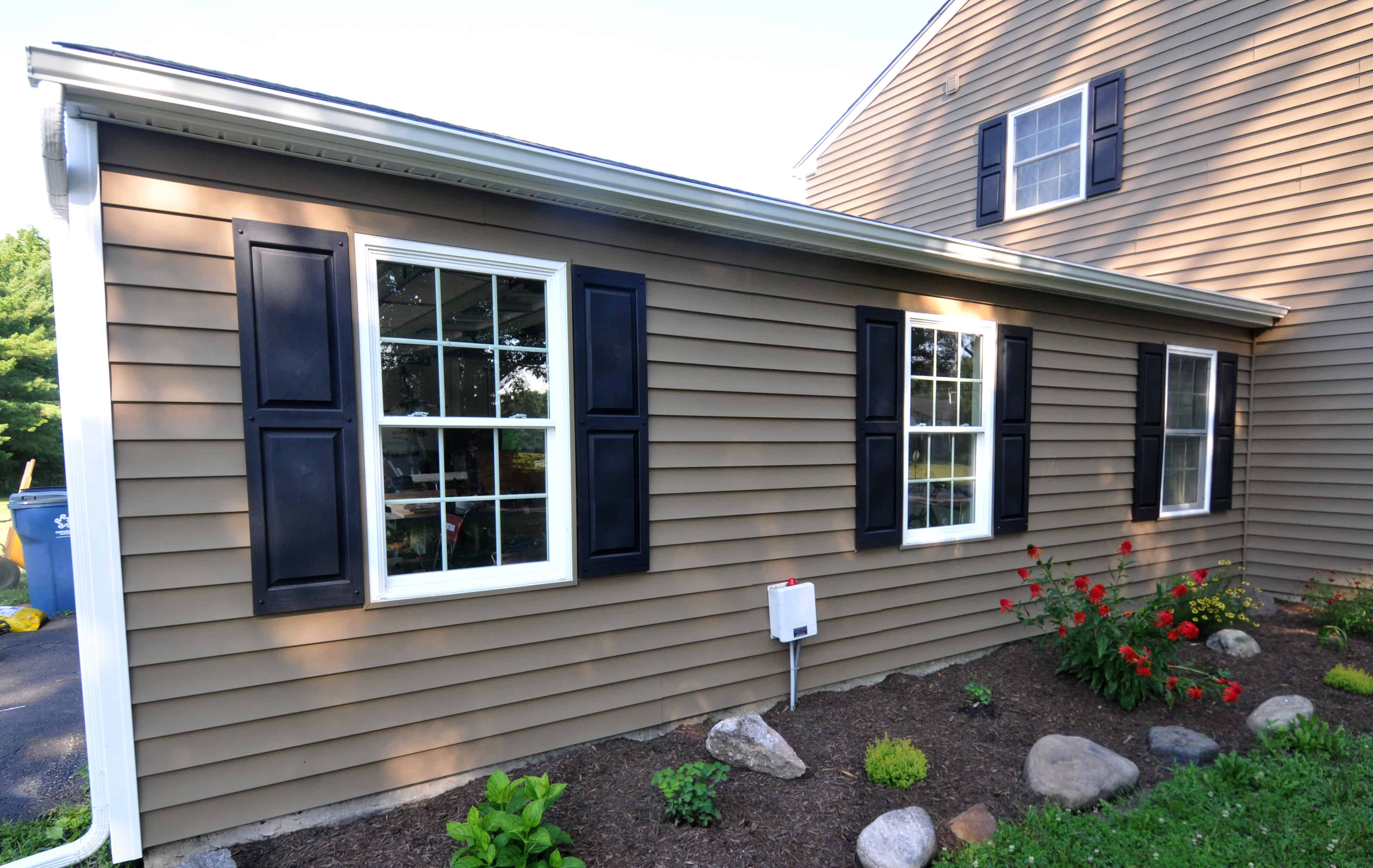 A brown house with white-trimmed windows and black shutters, featuring a small garden bed with rocks, green plants, and red flowers in front. A driveway and green trees are visible on the left side.