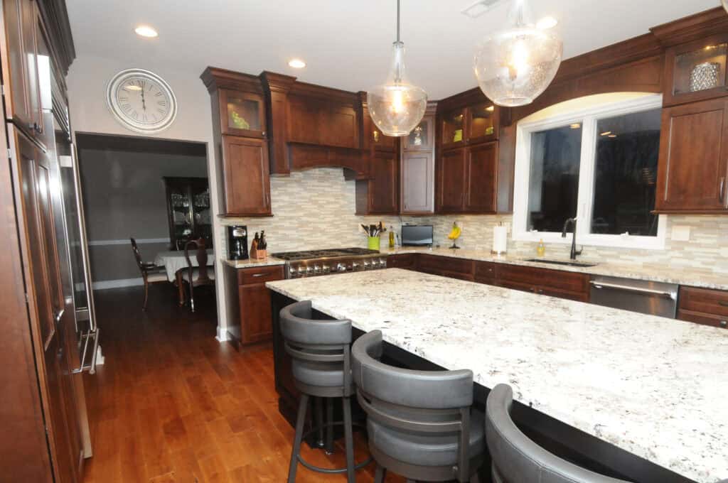 A modern kitchen with dark wood cabinets, granite countertops, stainless steel appliances, and four gray barstools at an island. Pendant lights hang above the island, and a dining area is visible in the background.