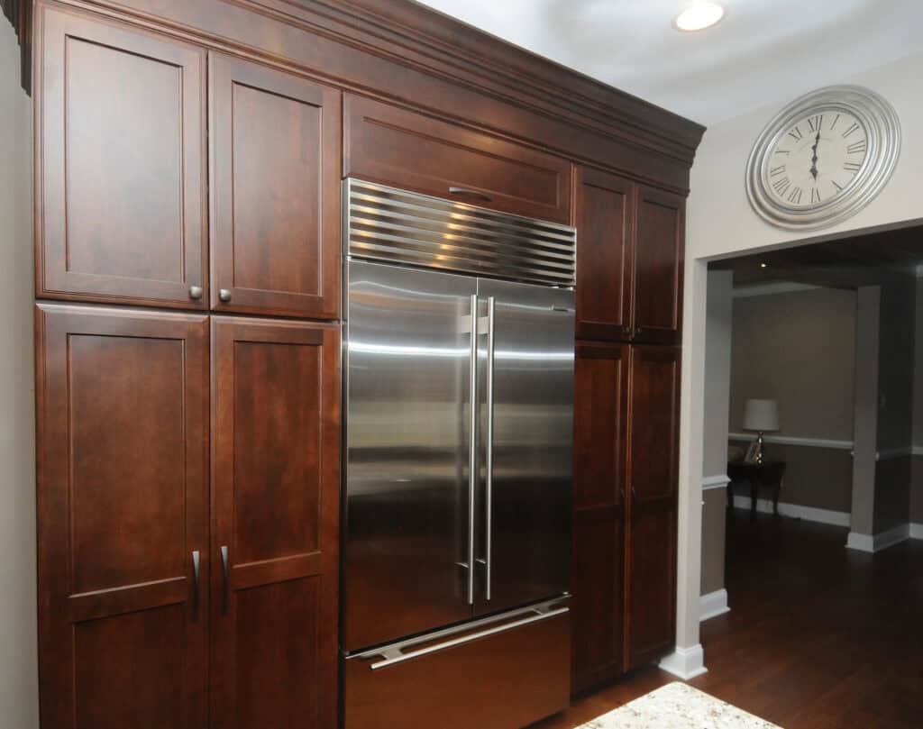Large stainless steel refrigerator with double doors, built into dark wooden kitchen cabinets. A round silver wall clock hangs above a hallway opening on the right. The kitchen has dark wood flooring and neutral walls.
