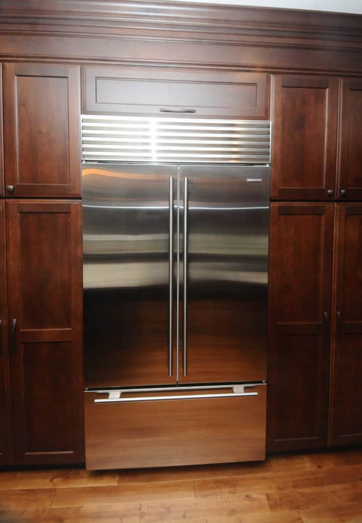 A stainless steel French door refrigerator with a bottom freezer is built into wooden kitchen cabinetry with dark brown paneled doors. The floor is hardwood.