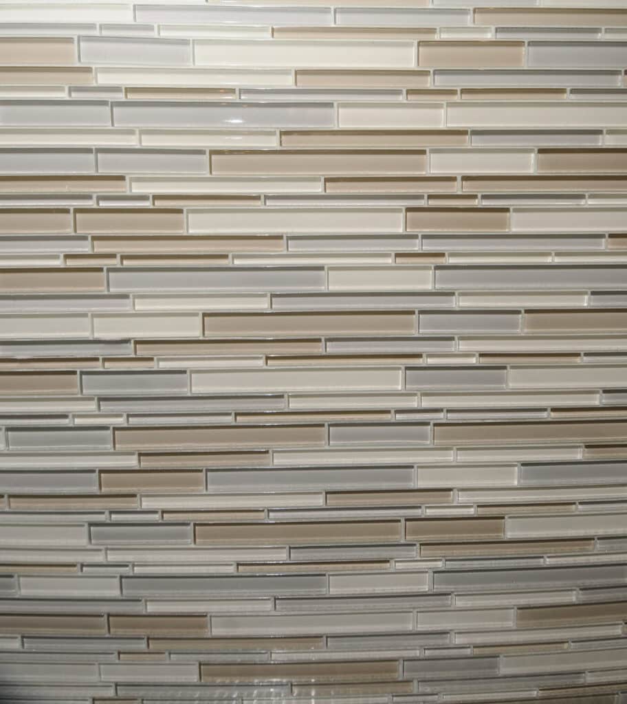 A close-up view of a mosaic tile wall with horizontal rectangular tiles in shades of beige, tan, cream, and light gray, arranged in a staggered pattern.
