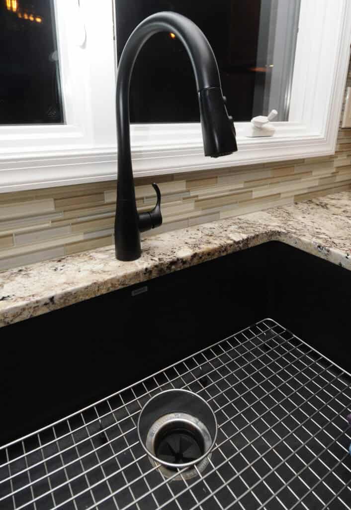 A modern black kitchen faucet over a large black sink with a metal grid at the bottom, set in a granite countertop in front of a window with a tiled backsplash.