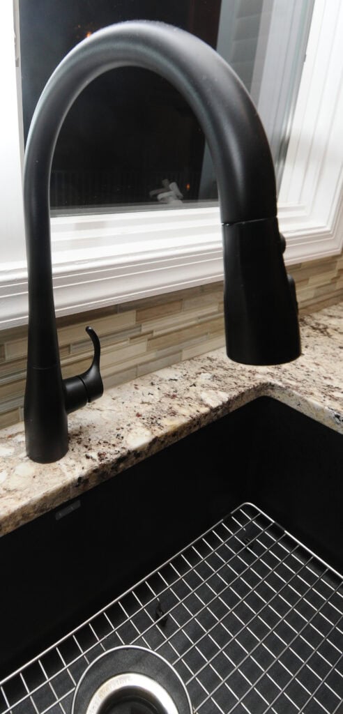 A matte black kitchen faucet with a curved spout is installed on a granite countertop next to a black sink with a metal rack, set against a tiled backsplash and a window.