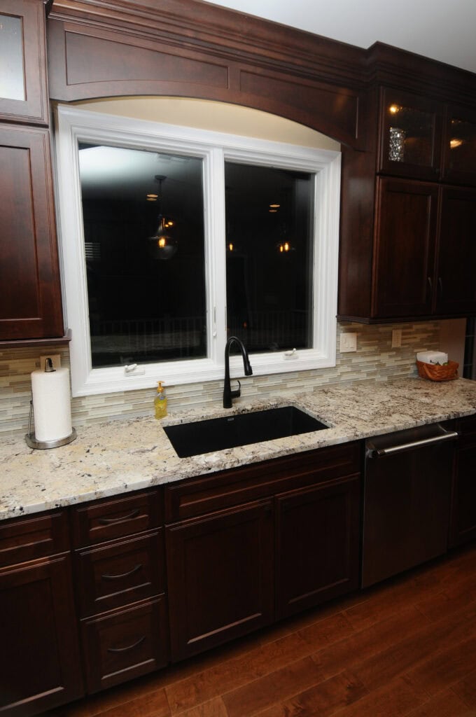 A modern kitchen sink with a black faucet is set in a granite countertop beneath a large window. Dark wood cabinets, a dishwasher, paper towel holder, soap dispenser, and a basket are visible.