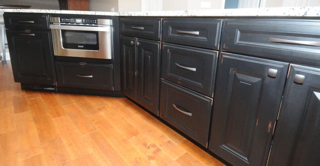 Row of dark wood kitchen cabinets with sleek handles, a built-in stainless steel microwave, and a granite countertop, all set on a light brown hardwood floor.
