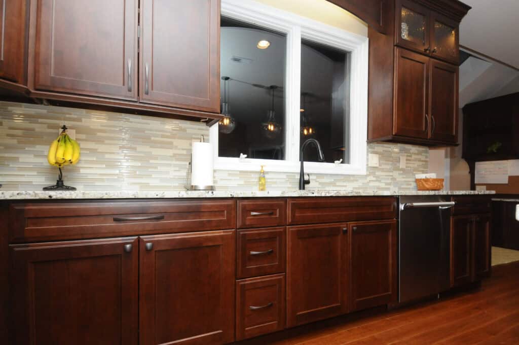 A modern kitchen with dark wood cabinets, a white and gray tiled backsplash, granite countertops, a sink under a large window, a dishwasher, and a banana holder on the counter.