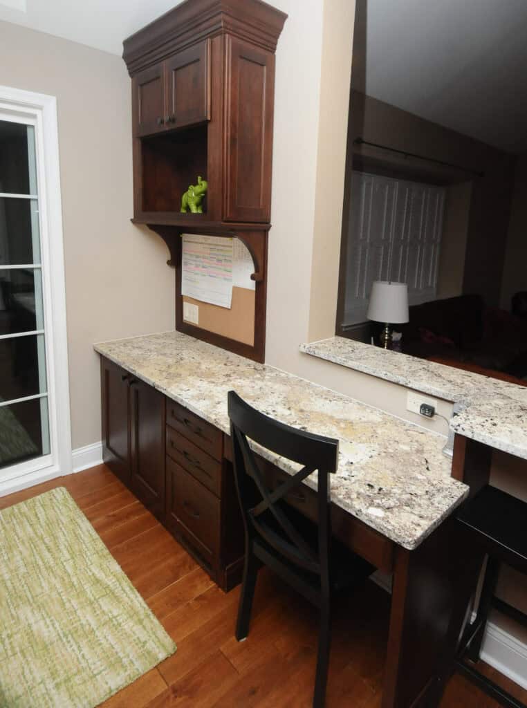 A home office nook with a granite countertop, dark wood cabinets, a black chair, a bulletin board with a calendar, and a green decorative object, next to a kitchen counter and window.