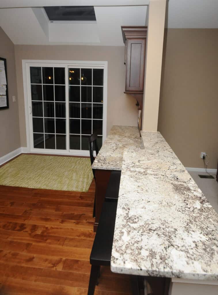 A modern kitchen with a granite countertop, two black bar chairs, light brown walls, wood flooring, a green rug, and large sliding glass doors leading outside.