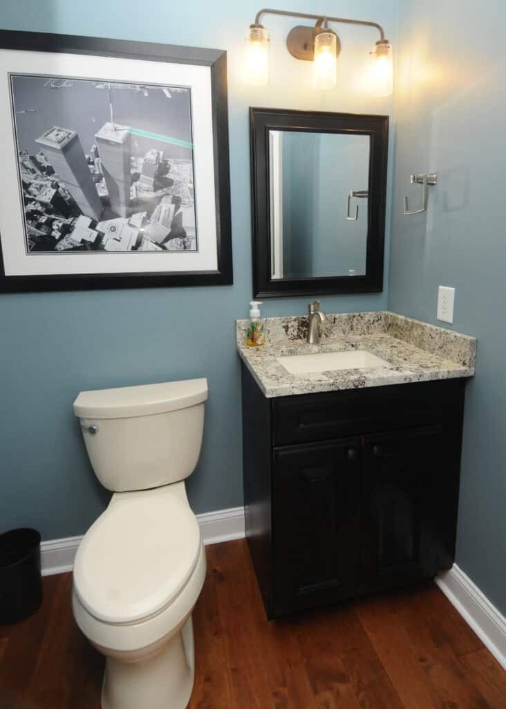 A small bathroom with blue walls, a white toilet, a black vanity with a granite countertop and sink, a rectangular mirror, wall-mounted light fixture, and a framed black-and-white cityscape photo above the toilet.