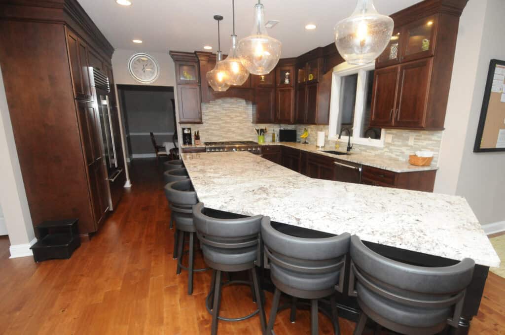 Spacious kitchen with dark wood cabinets, a large marble island with six black barstools, stainless steel appliances, pendant lights, and wood flooring. A window and wall clock are also visible.