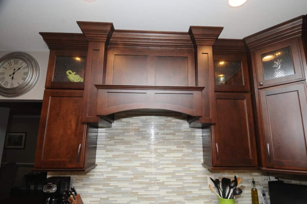 Dark wood kitchen cabinets with decorative molding, glass-front upper sections, and a neutral-toned tile backsplash. A wall clock and various kitchen utensils are visible in the scene.