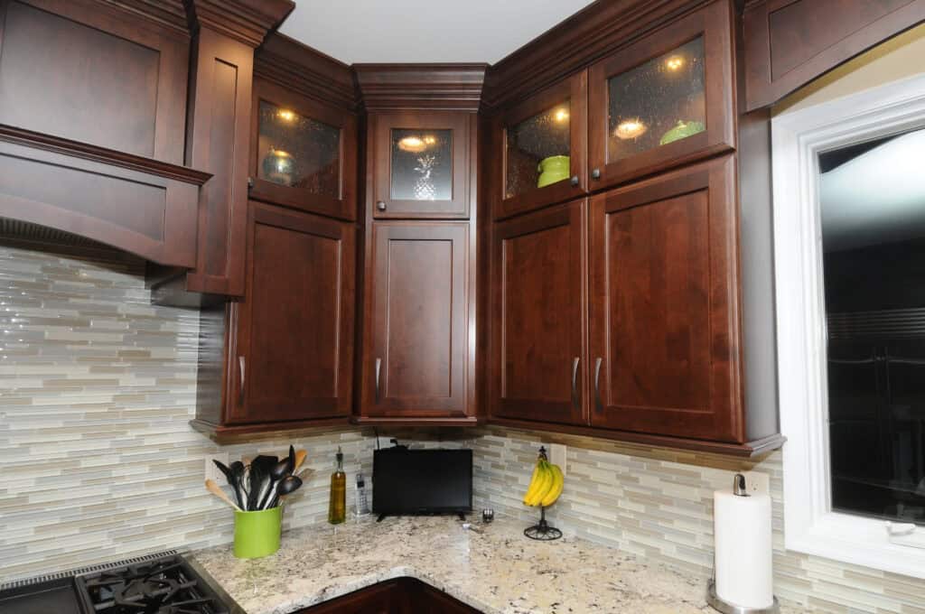 A kitchen corner with dark wood cabinets, some with glass panels showing green dishes inside. Granite countertop holds utensils in a green holder, olive oil, bananas, a paper towel roll, and a small black screen.