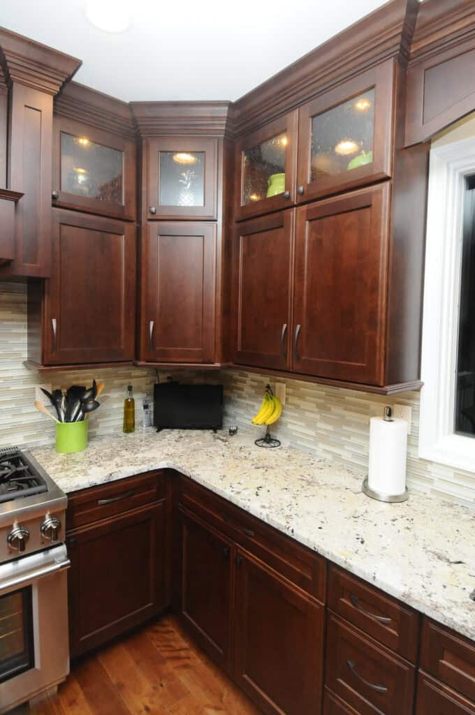Modern kitchen with dark wood cabinets, granite countertops, a gas stove, knife block, olive oil bottle, bananas, paper towel holder, and a small appliance on the counter. Light tiled backsplash and wooden floor.