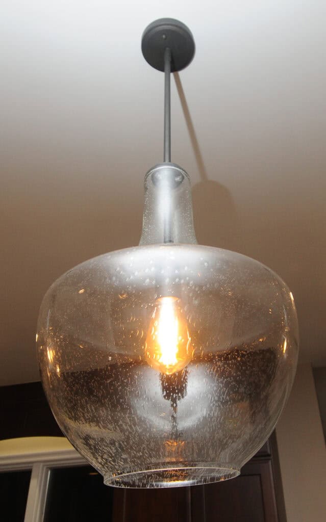 A close-up view of a hanging glass pendant light fixture with a visible glowing bulb inside, suspended from a ceiling. The glass shade has a slightly bubbled texture.