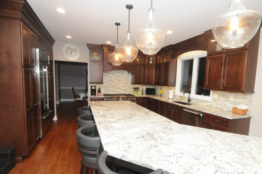 A modern kitchen with dark wood cabinets, stainless steel appliances, a large white and gray granite island with black barstools, pendant lighting, and a wall clock above the entryway.