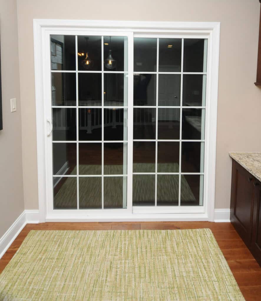 A glass sliding door with white grid frames, leading to a dark area. The interior features light-colored walls, a beige and green rug on wooden flooring, and a granite countertop on the right.