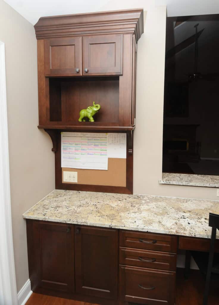 A kitchen nook with dark wood cabinets, granite countertop, a corkboard with a calendar pinned on it, and a decorative green elephant figurine placed on a shelf above the counter.