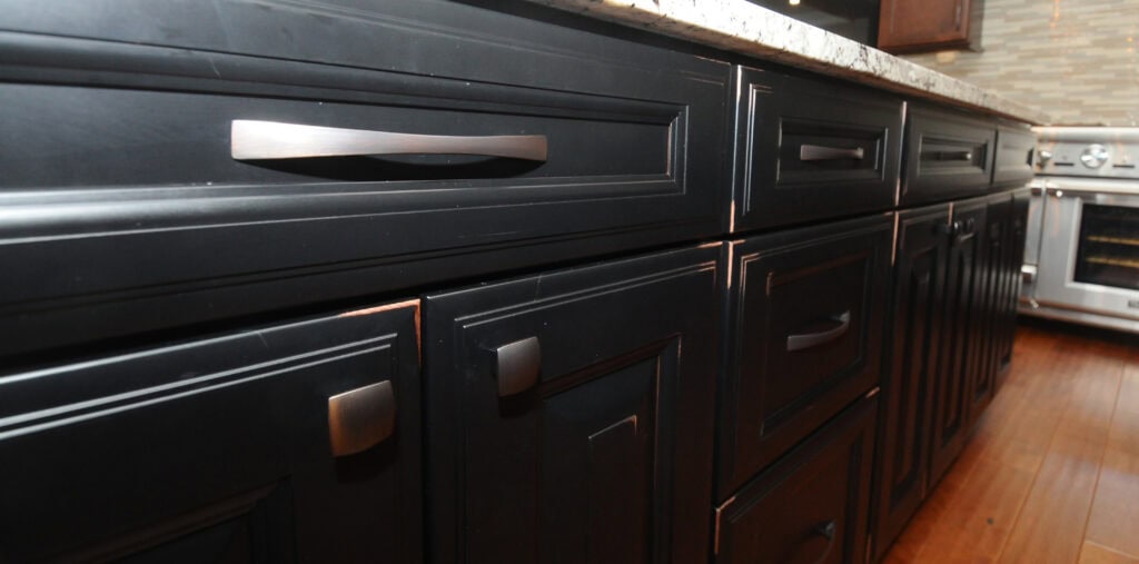 Close-up of black kitchen cabinets and drawers with modern metal handles, a granite countertop above, and a stainless steel oven visible in the background. The floor is wooden.