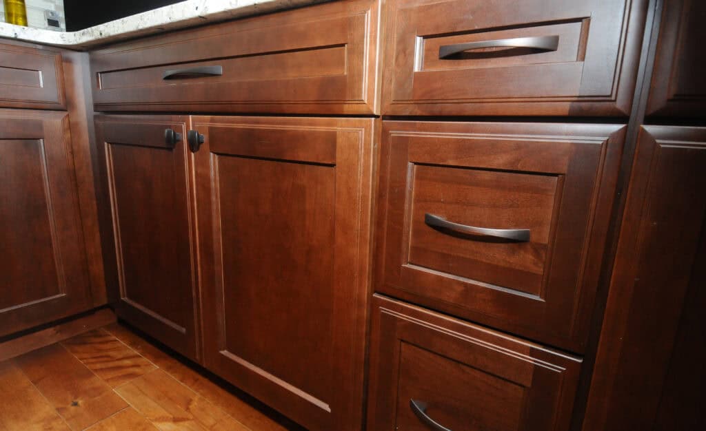 Close-up of dark wooden kitchen cabinets and drawers with black handles, set beneath a light-colored countertop and placed on a matching wooden floor.