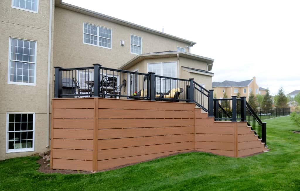 A modern two-story house with a raised wooden deck featuring black railings, outdoor furniture, and stairs leading down to a well-manicured green lawn. Neighboring houses are visible in the background.