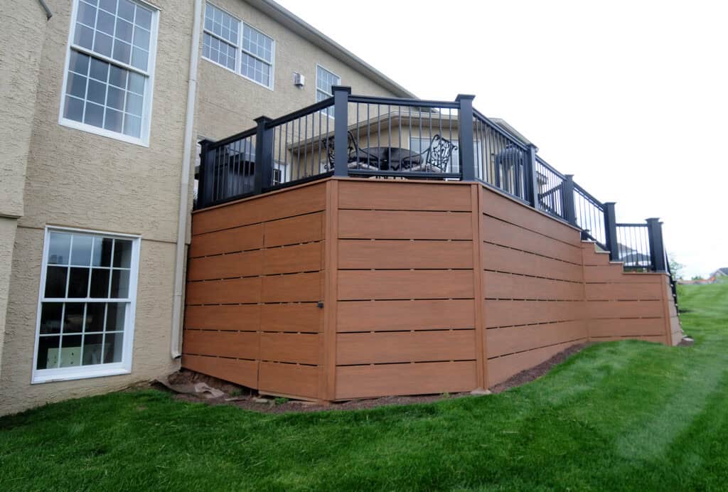 A modern raised deck with black railings and horizontal wooden panels below, attached to a beige house. The deck overlooks a green, neatly mowed lawn on a sloped yard.