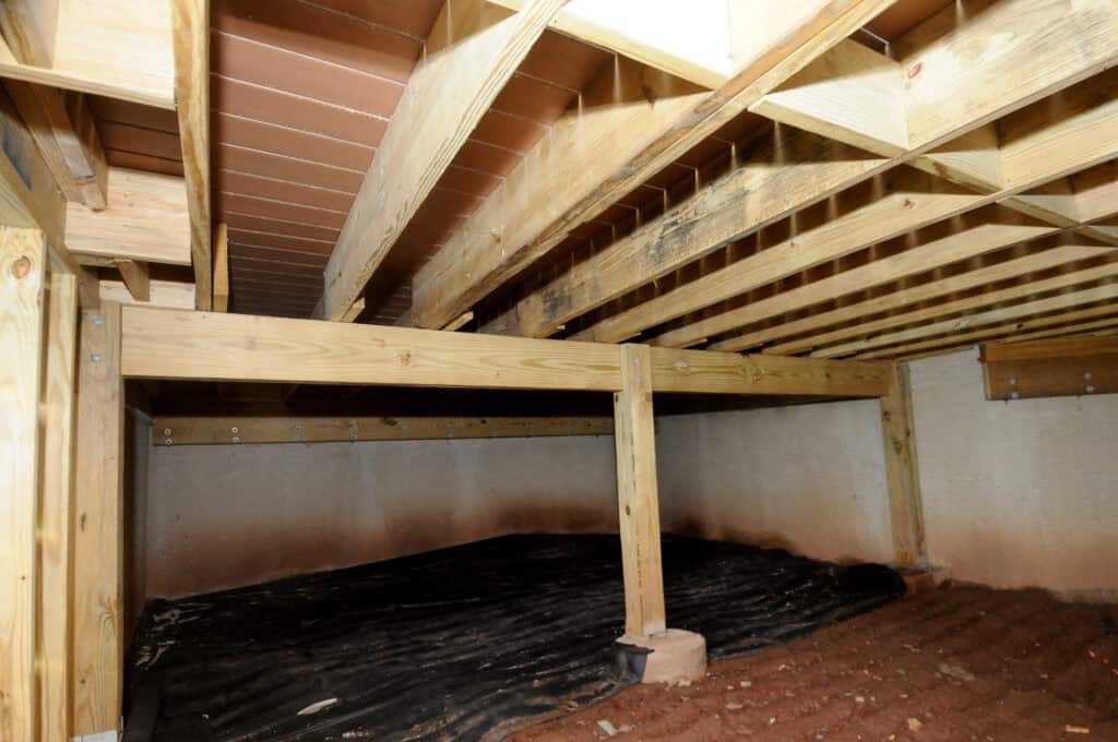 A crawl space with exposed wooden beams and support posts, dirt floor partly covered with black plastic sheeting, and concrete or cinder block walls.