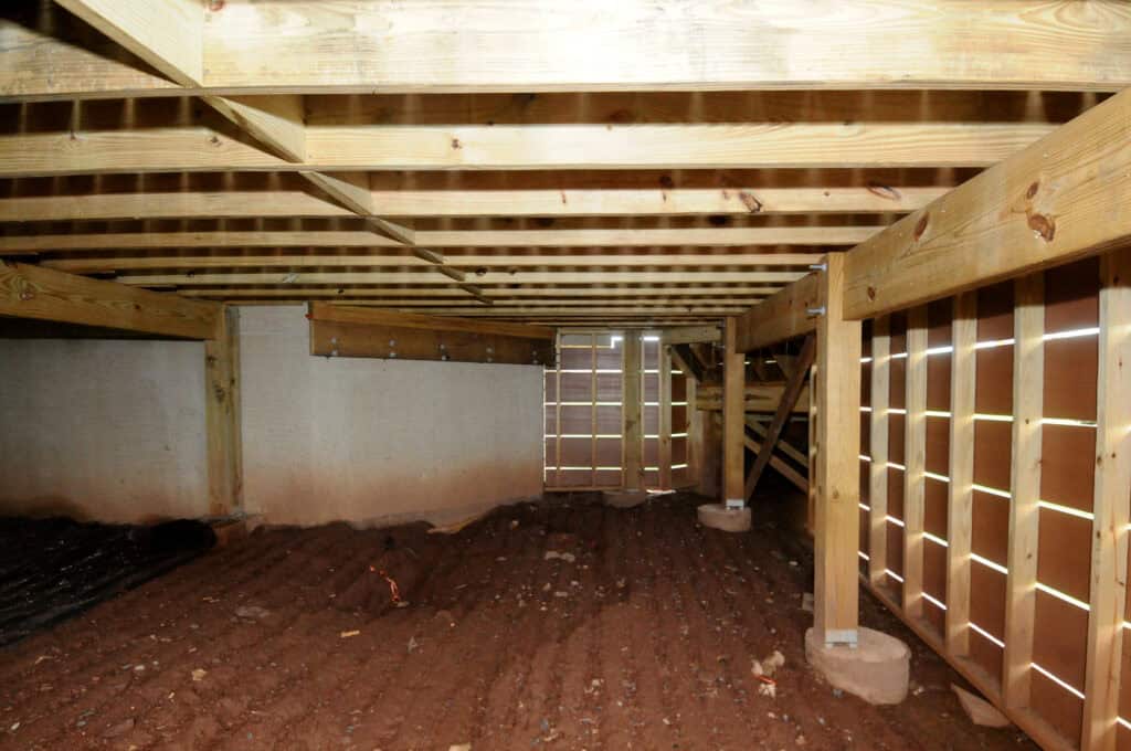 A clean, well-lit crawl space under a house with exposed wooden beams, a dirt floor, and partial walls made of wood and concrete. Light filters through slats in the wooden structure.