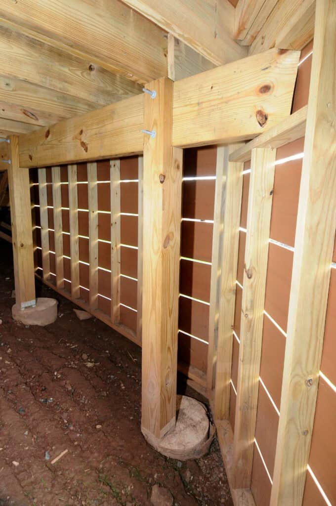 Wooden support beams and vertical posts form part of an under-house structure, with a slatted wooden wall allowing light through. The ground below is bare, uneven soil.