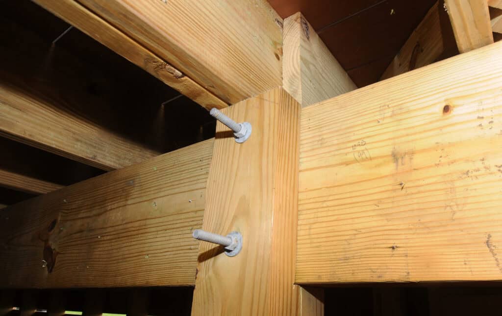 Close-up of a wooden deck structure showing two large bolts securing intersecting beams and posts, demonstrating construction details and connection points under the deck.