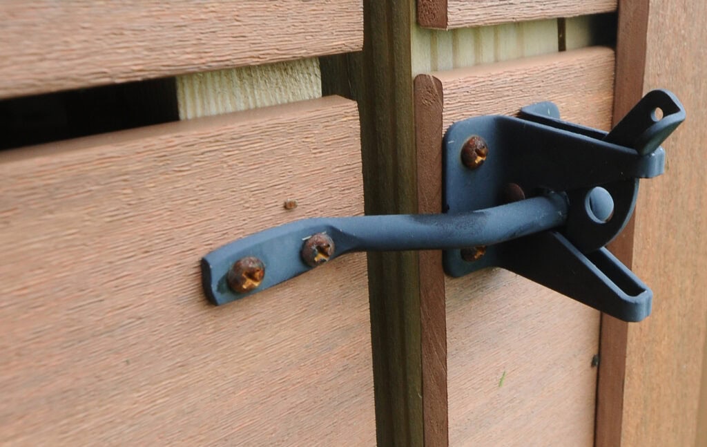 A close-up of a black metal latch secured to two wooden panels, with some visible rust on the screws. The latch holds the panels together and the wood has a reddish-brown finish.