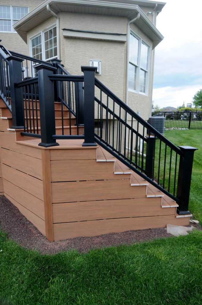 A modern outdoor staircase with light brown wooden steps and black metal railings, attached to the side of a beige house with large windows, leading down to a green lawn.