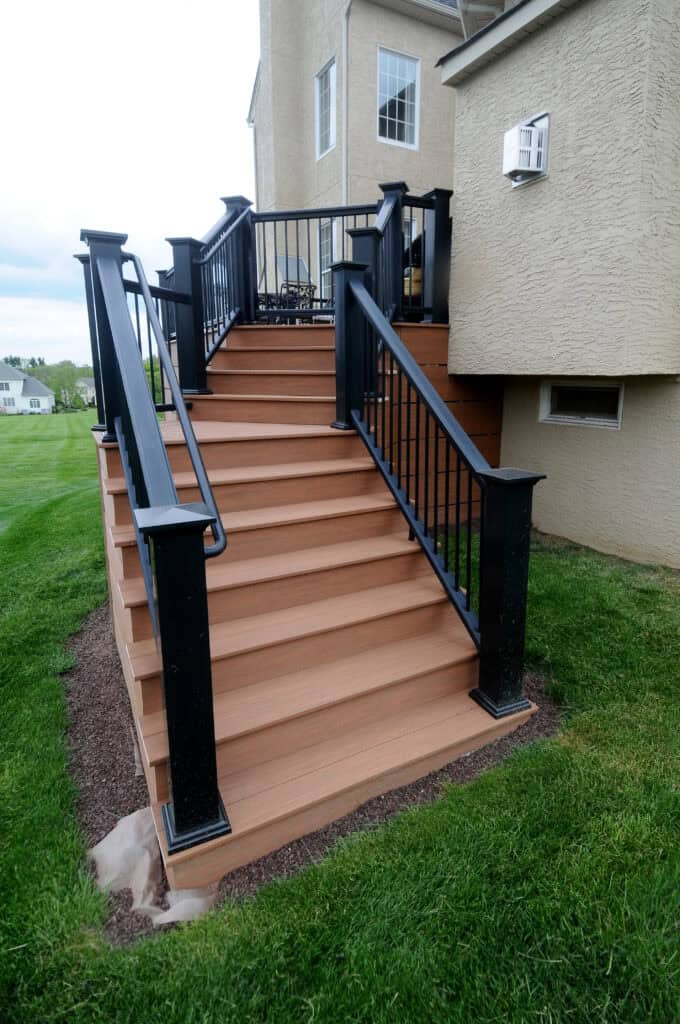 A set of brown wooden stairs with black railings leads from a raised deck to a grassy yard next to a beige house. The stairs have black metal balusters and posts.
