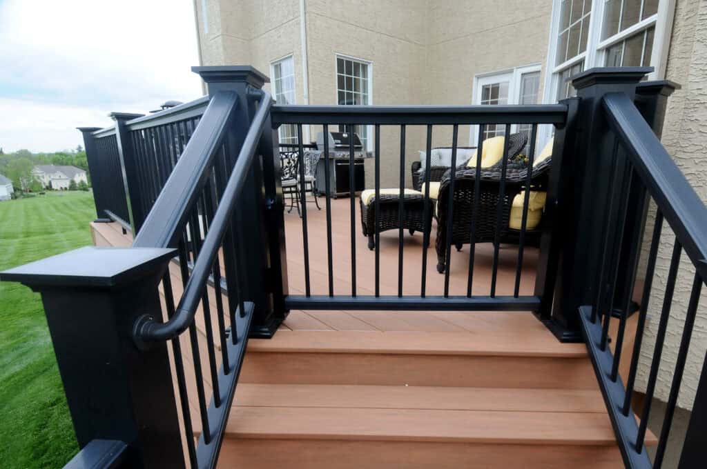 A set of brown wooden steps with black railings leads up to a patio deck with wicker chairs, yellow cushions, and a grill next to a beige house with large windows.