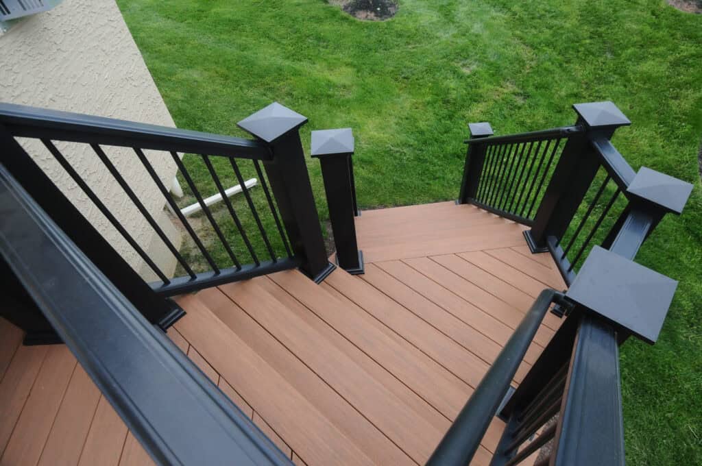 A brown wooden deck staircase with black metal railings and decorative posts leads down to a green grassy yard next to a beige textured house wall.