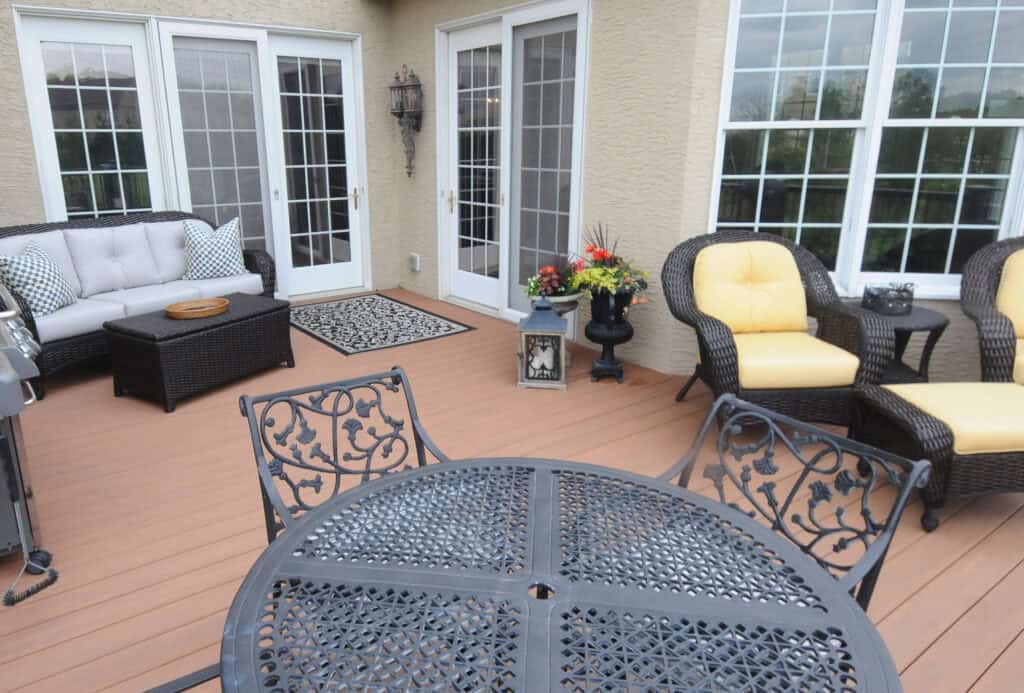 A patio with brown wooden flooring, black metal table and chairs, wicker sofa with pillows, wicker armchairs with yellow cushions, a small rug, potted flowers, and large glass doors and windows.