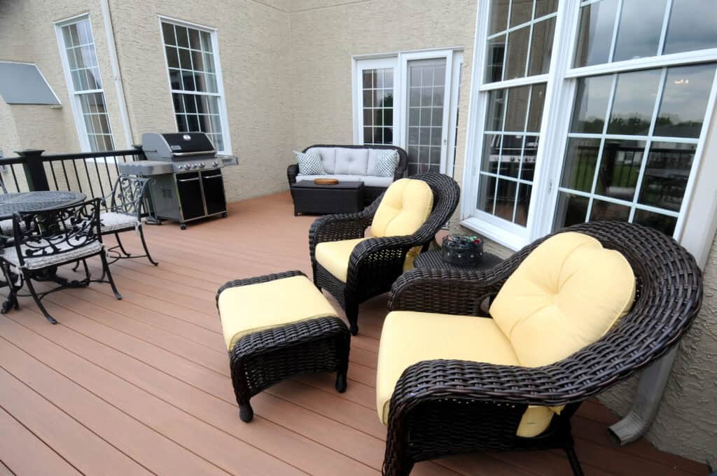 A spacious outdoor patio features wicker chairs with yellow cushions, a matching ottoman, a metal dining set, a grill, and a loveseat with neutral cushions, all set against a beige house with large windows and glass doors.