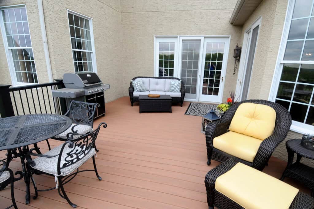 A spacious patio with wicker furniture, including chairs with yellow and white cushions, a metal table set, a barbecue grill, and a small sofa, set against a beige house with large windows and glass doors.
