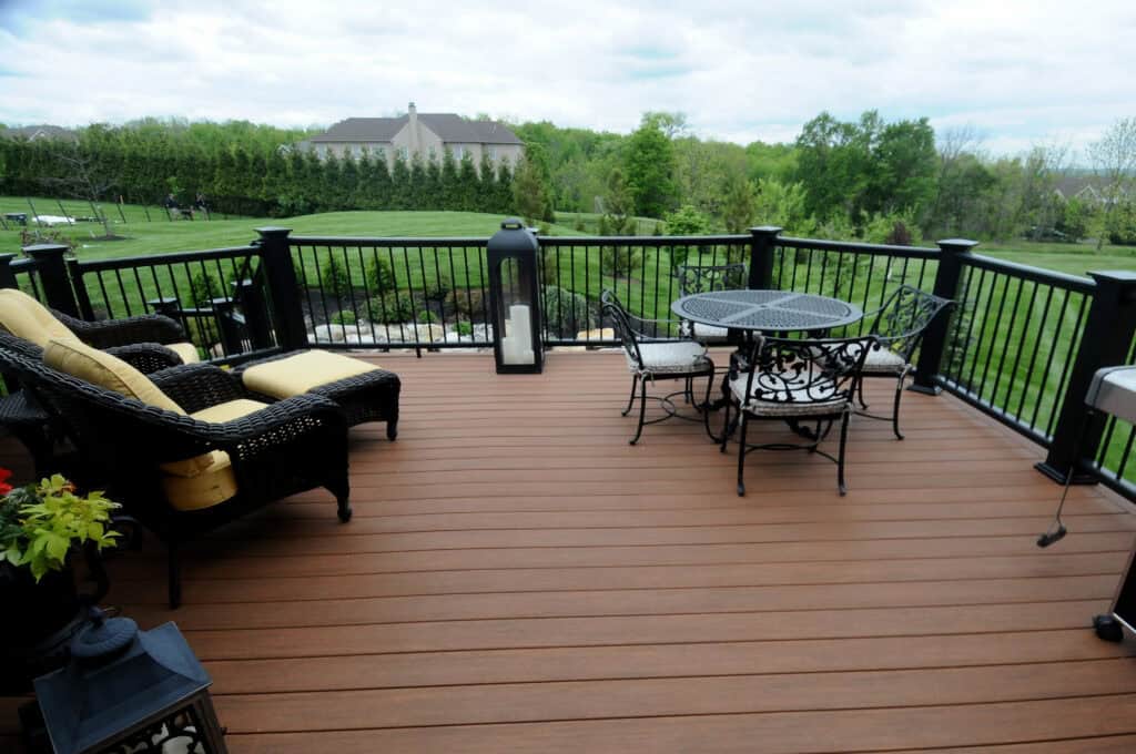 A spacious backyard deck with wicker seating, a round metal table and chairs, black railings, and a scenic view of green lawn, trees, and a house in the distance under a cloudy sky.