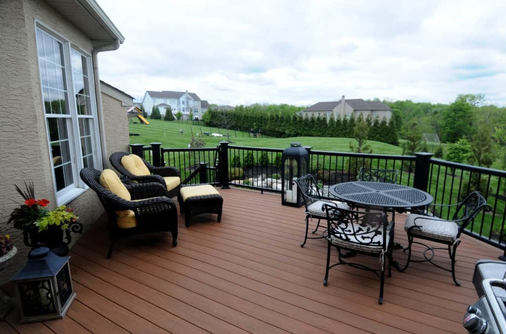 A backyard deck with wicker chairs, yellow cushions, a metal table and chairs, a grill, and potted plants, overlooking a large green lawn with neighboring houses and trees in the background.