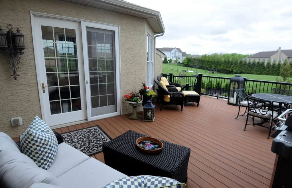 A cozy wooden patio with cushioned seating, a small table, potted plants, and decorative pillows, overlooking a green backyard and neighboring houses under a cloudy sky.