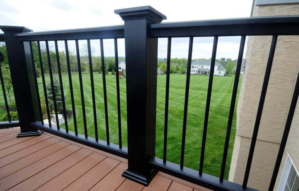 View from a deck with brown wooden flooring and a black metal railing, overlooking a large, neatly mowed green lawn with trees and houses in the distance under a cloudy sky.