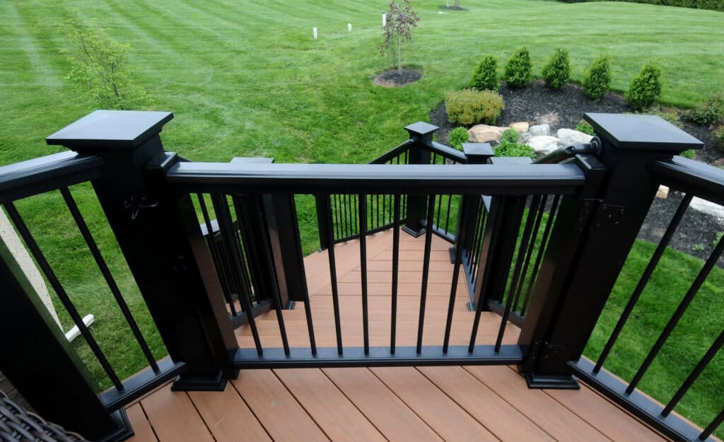 View from a deck with black railings and brown steps leading down to a landscaped backyard with neatly mowed green grass, small trees, and shrubs.