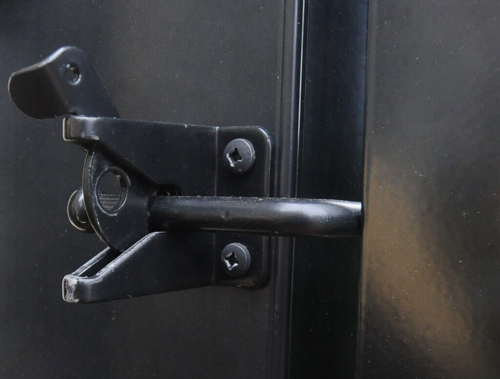 Close-up of a black latch lock fastened to a dark metal door, with screws visible securing the latch mechanism in place.