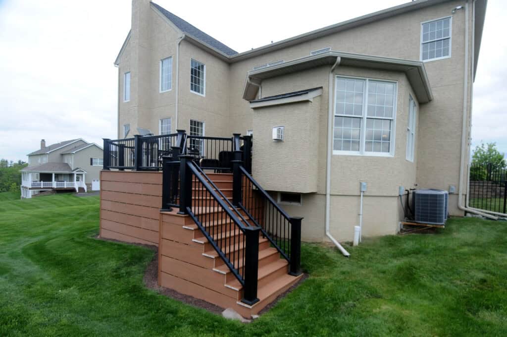 Two-story beige house with a raised wooden deck featuring black railings and stairs leading down to a well-kept green lawn. An air conditioning unit is visible beside the house.