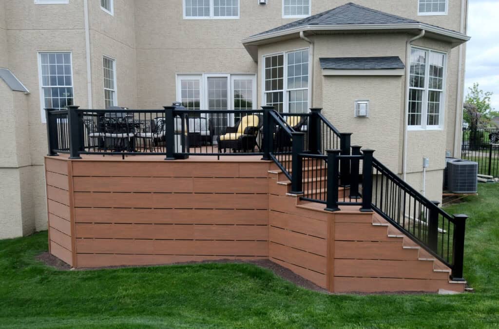 A raised wooden deck with black railings and stairs, attached to the back of a beige house with large windows, overlooking a green grassy yard. Outdoor seating is visible on the deck.