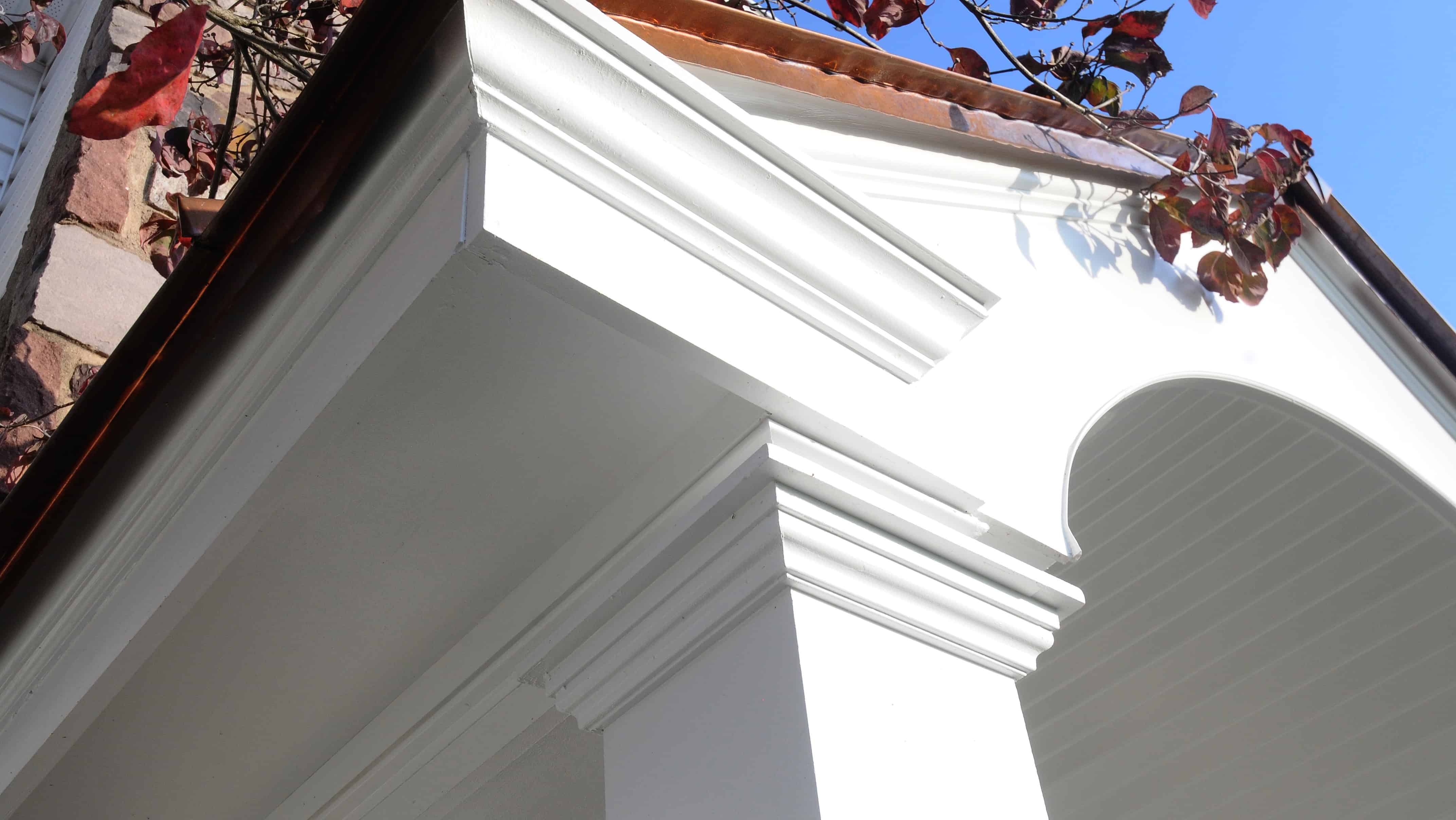 Close-up view of white architectural molding and columns on a house porch, with a brown copper roof edge and red leaves visible against a clear blue sky.